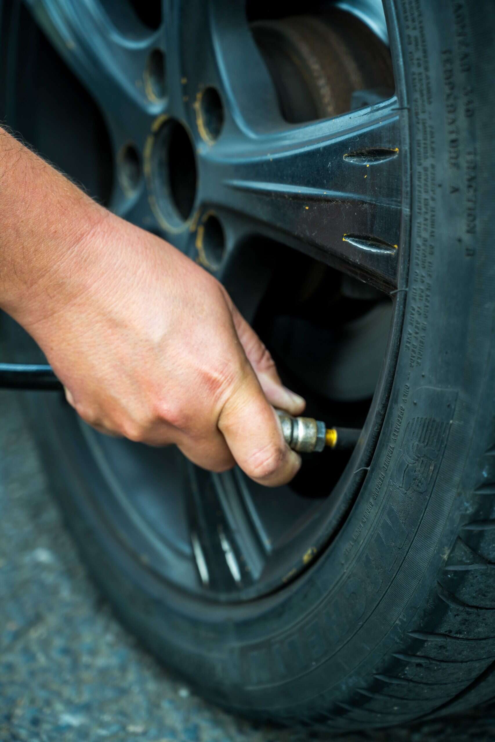 Checking Tire Pressure on a Passenger Vehicle Close-up of a person's hand using an air pressure gauge to check a car tire's valve stem, with the tire and wheel clearly visible on a paved surface.